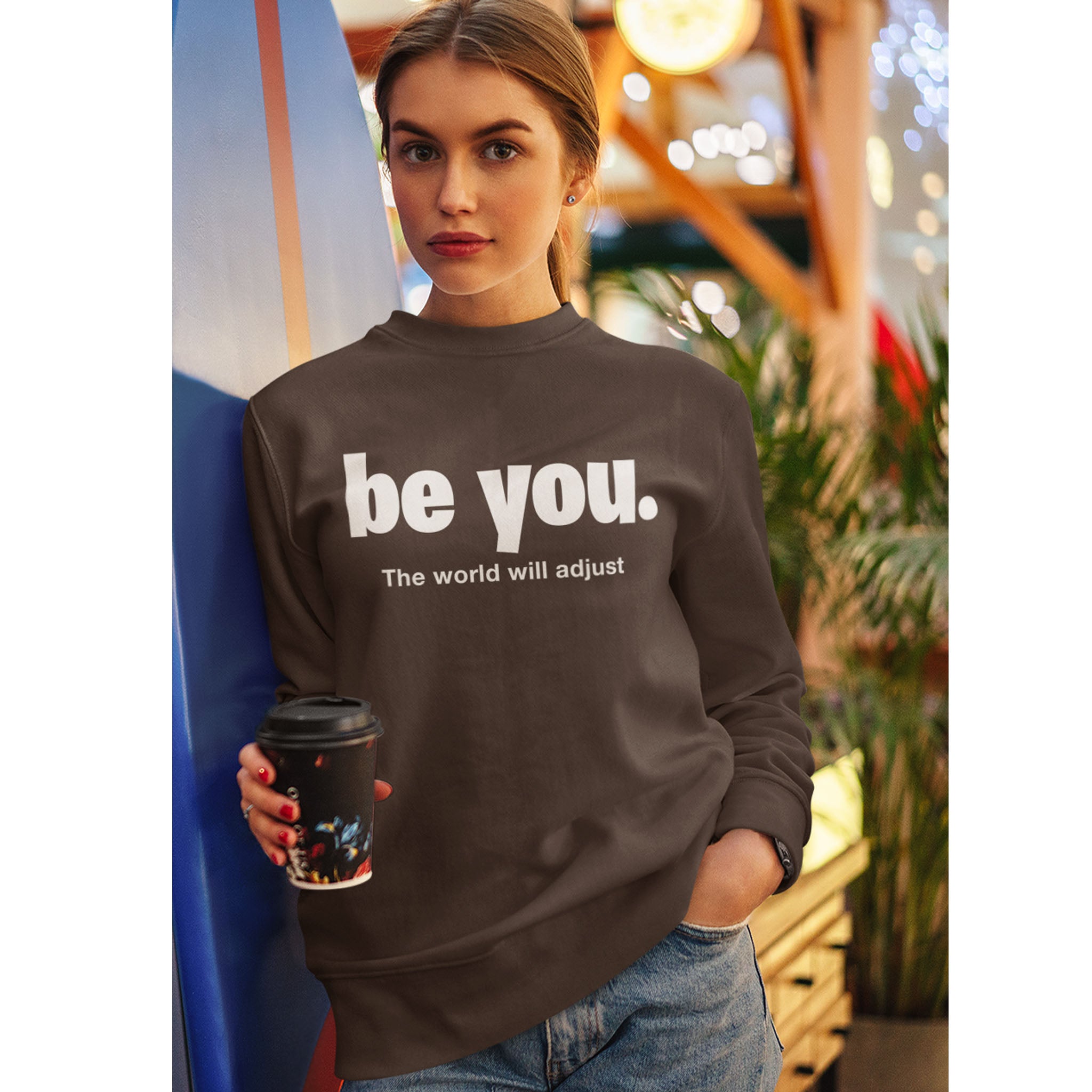 Person wearing a brown sweatshirt with 'be you. The world will adjust' text, holding a coffee cup.