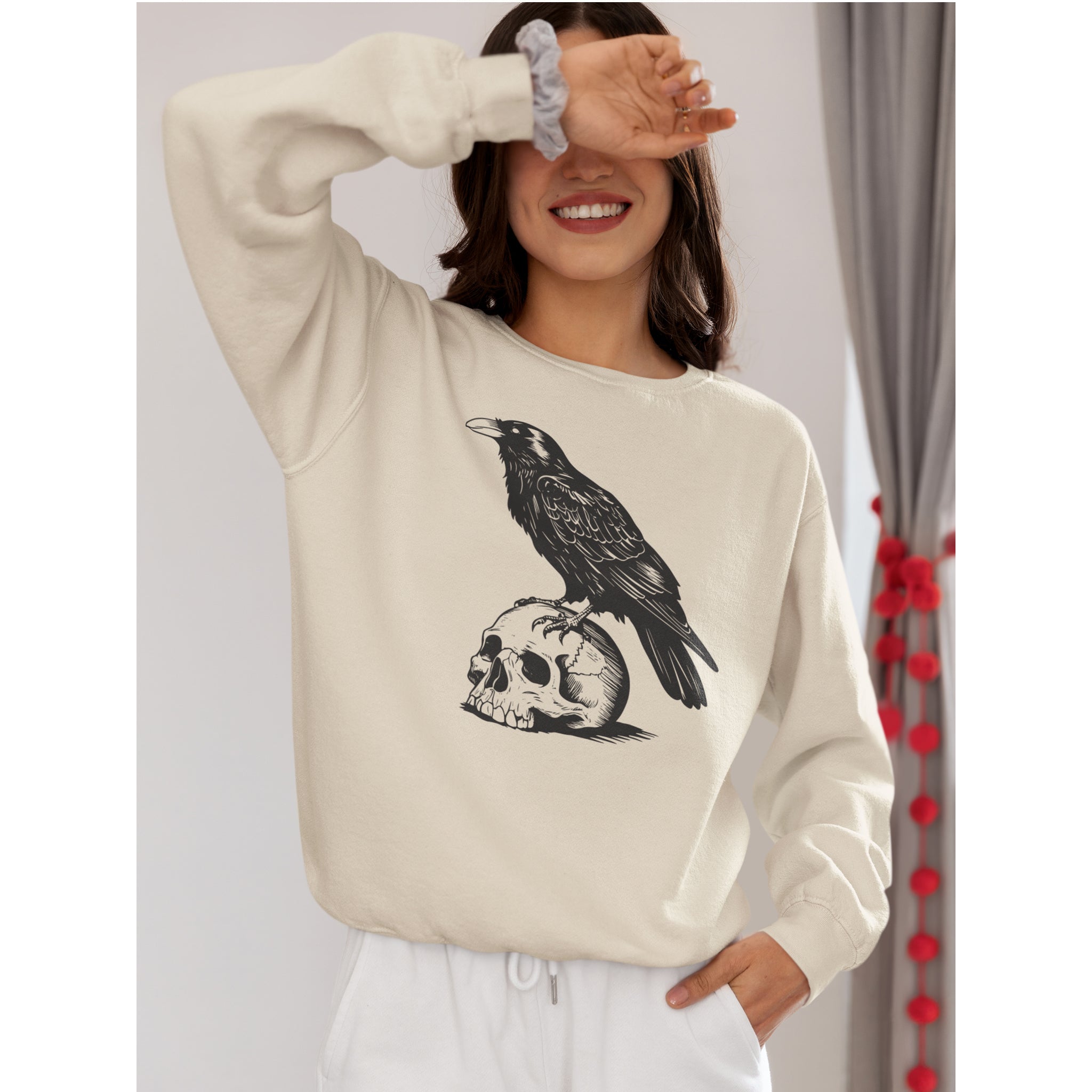 Person wearing a beige sweatshirt with a graphic design of a crow on a skull, standing against a plain background.