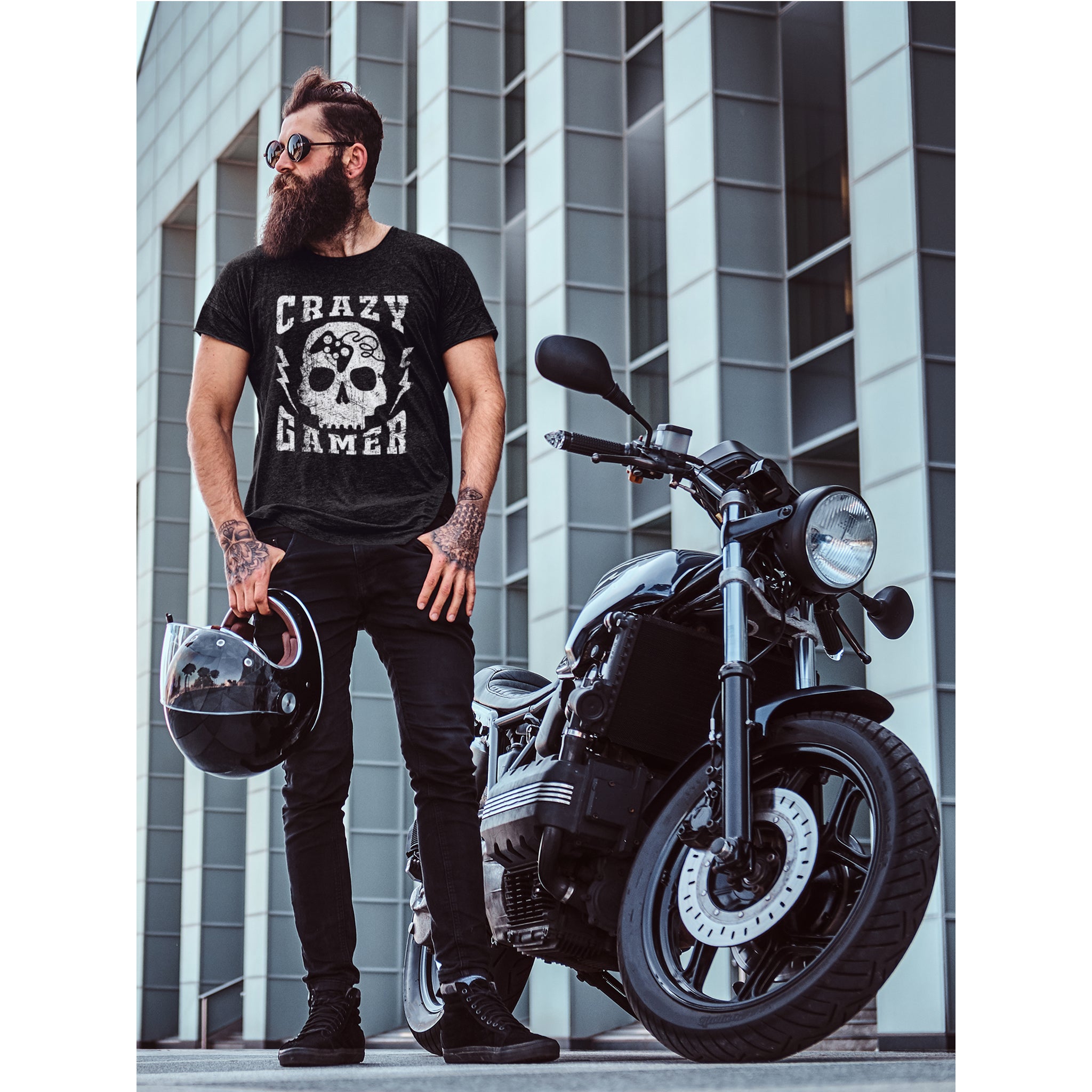 Man wearing a 'Crazy Gamer' t-shirt standing next to a black motorcycle with a modern building in the background.