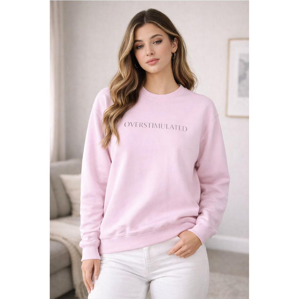 Woman wearing a pink sweatshirt with 'OVERSTIMULATED' text in a living room setting.