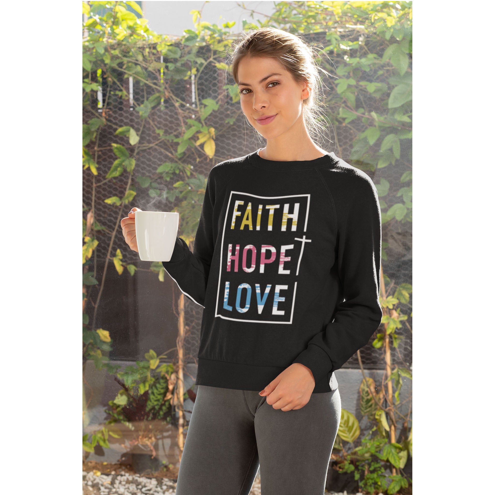 Woman wearing a black sweatshirt with 'Faith Hope Love' text, holding a white mug outdoors.