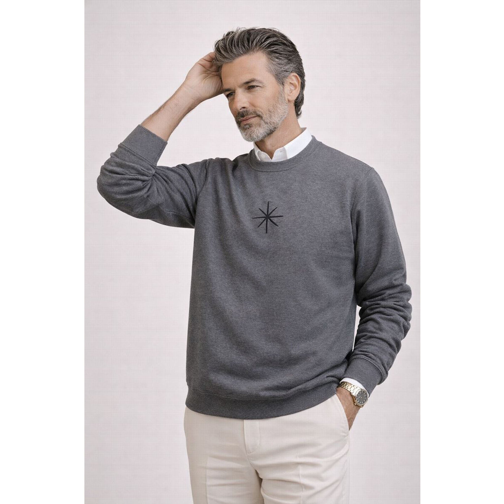 Man wearing a gray sweater with a logo, white shirt, and beige pants on a light background