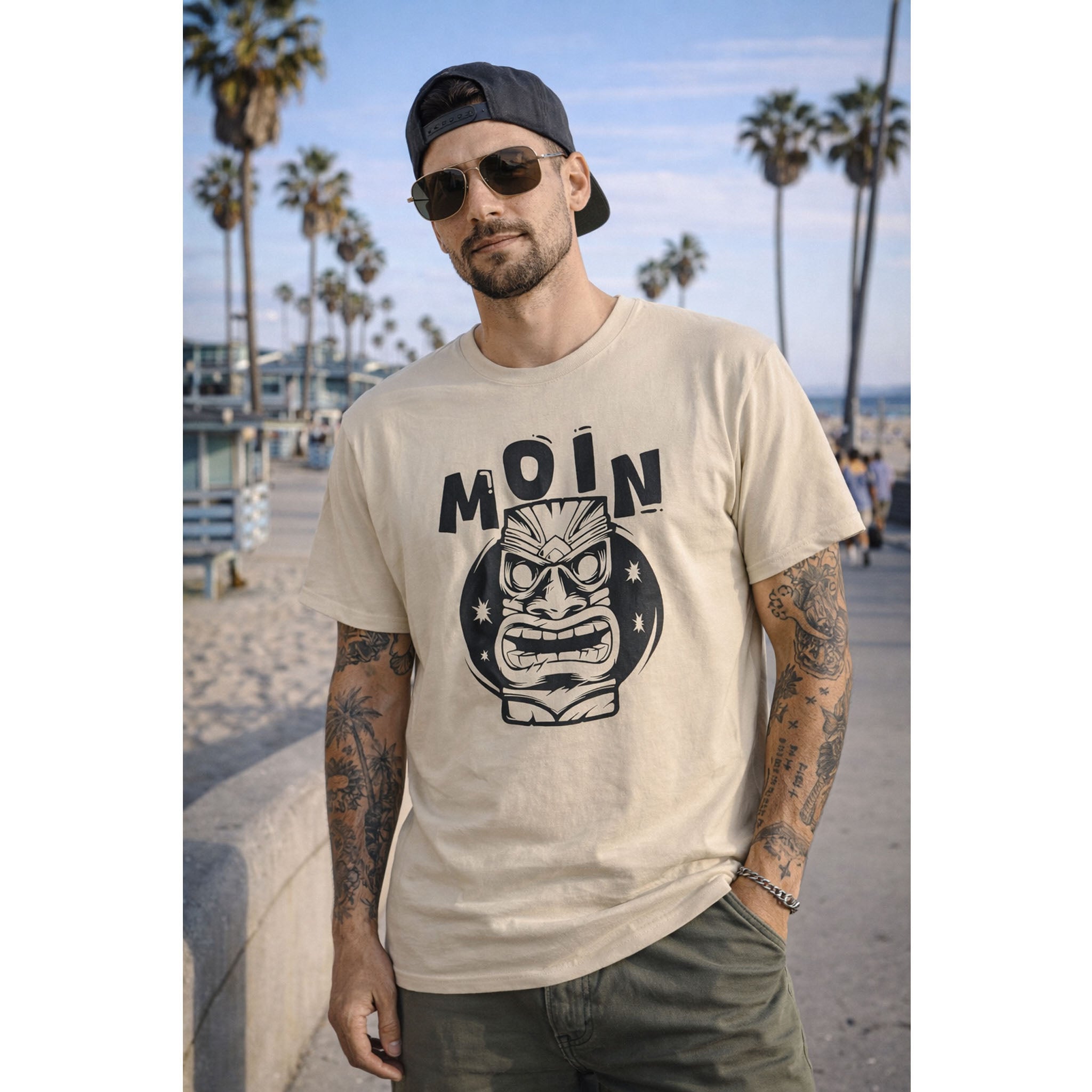 Man wearing a beige t-shirt with a graphic design and text, standing on a beach with palm trees in the background – Bamerix