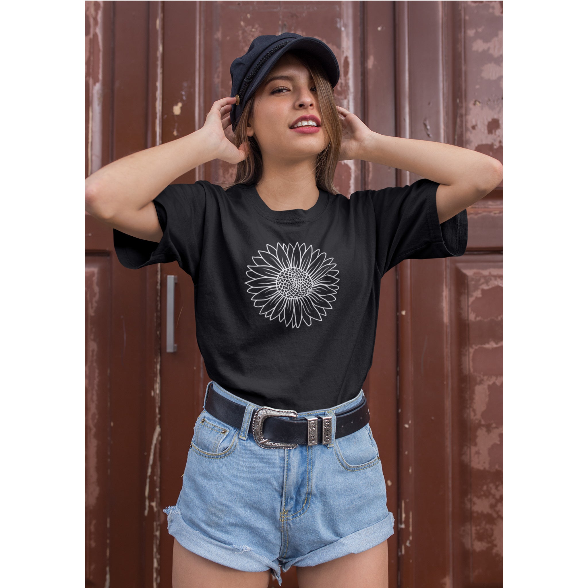 Woman wearing a black t-shirt with a sunflower design, blue shorts, and a cap against a brown door background.