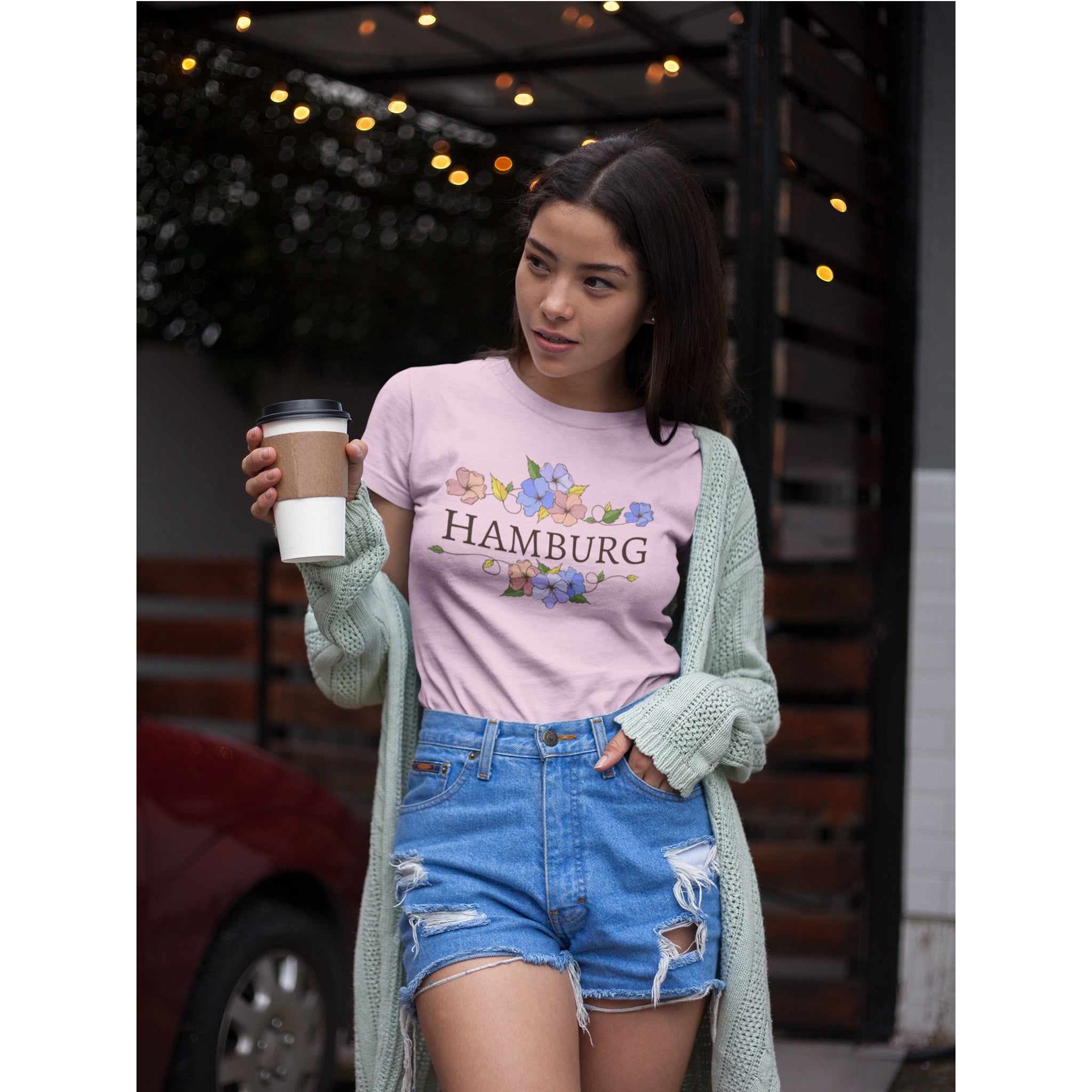 Woman wearing a pink t-shirt with 'Hamburg' and floral designs, holding a coffee cup.