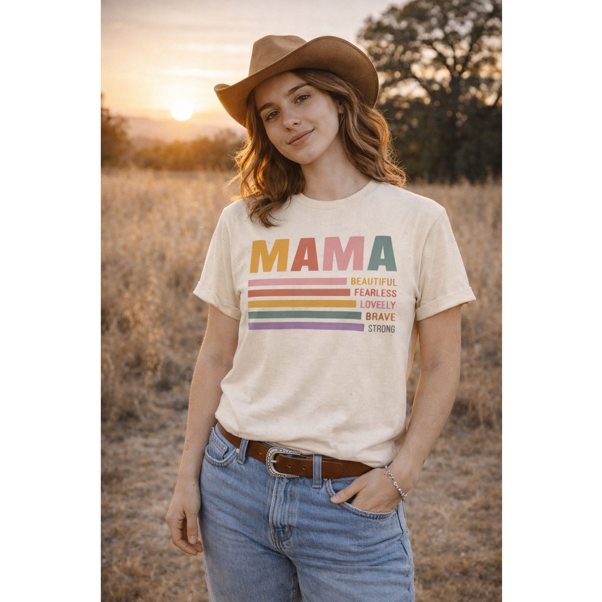 Woman wearing a 'MAMA' t-shirt with colorful text in a field at sunset