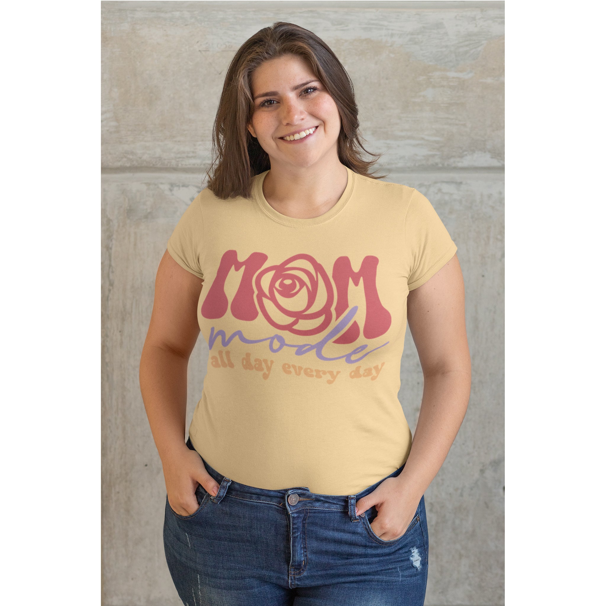 Woman wearing a beige t-shirt with 'Mom mode all day every day' text against a concrete wall.