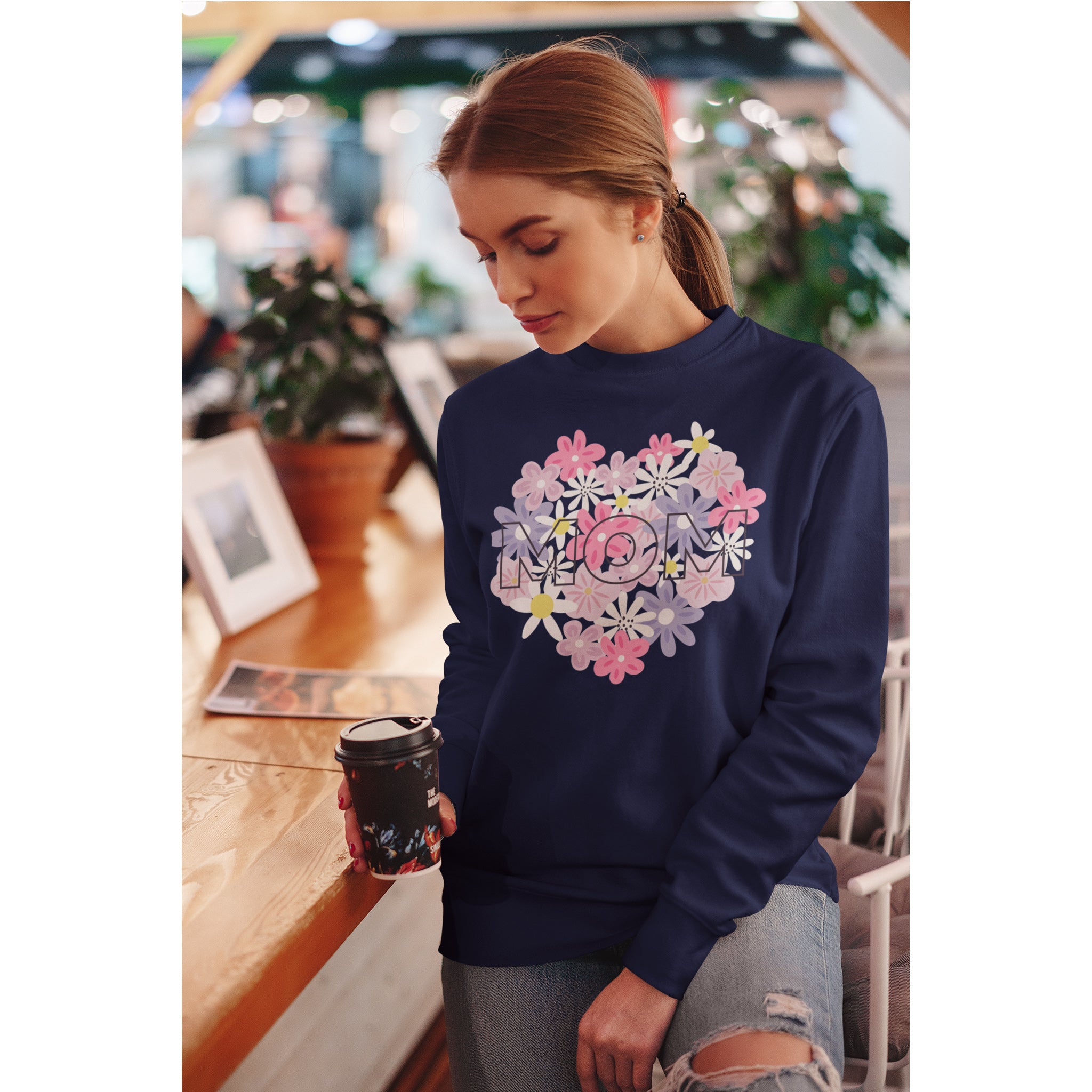 Woman wearing a navy sweatshirt with floral 'MOM' design in a cafe.