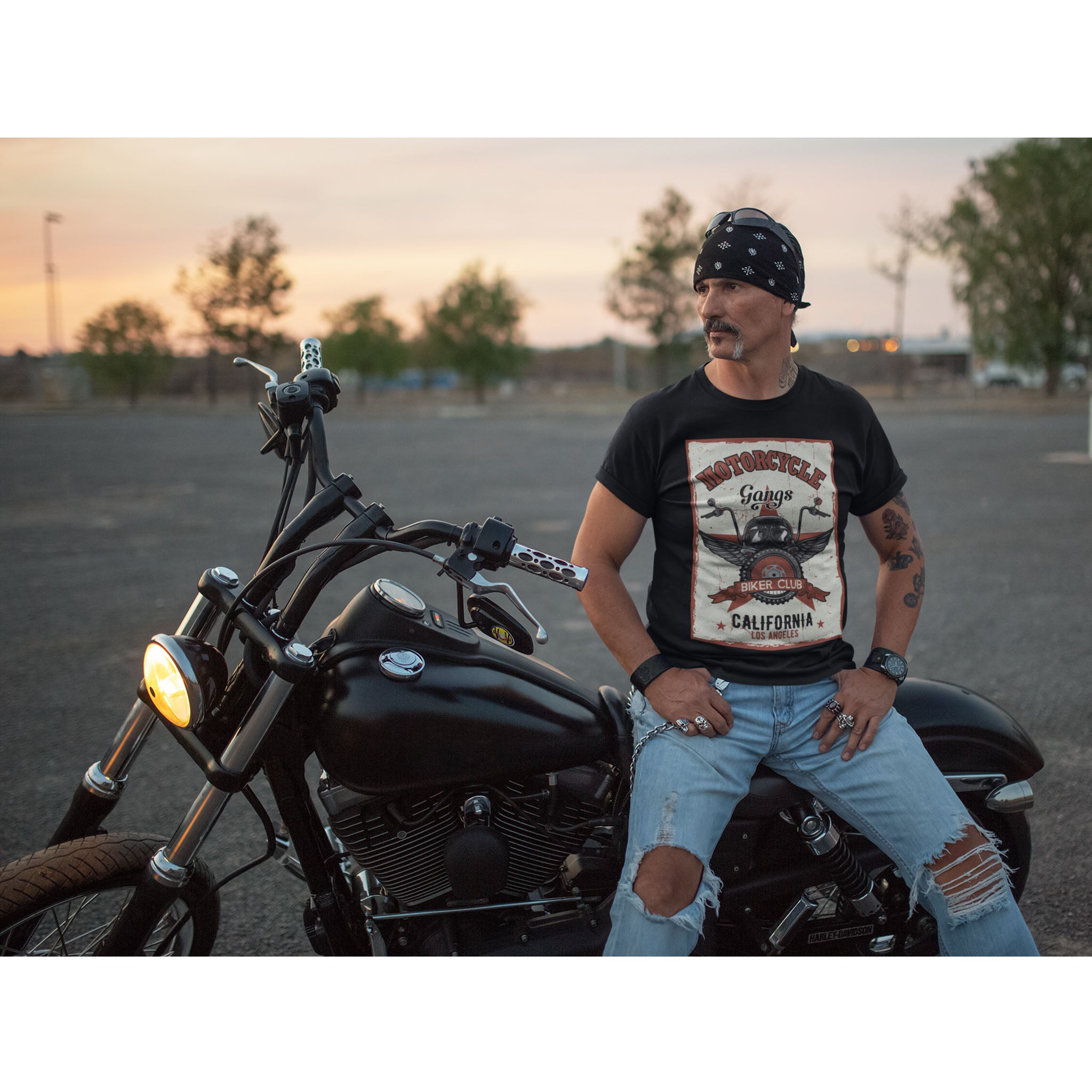 Man sitting on a motorcycle wearing a bandana and casual clothing with a sunset background.