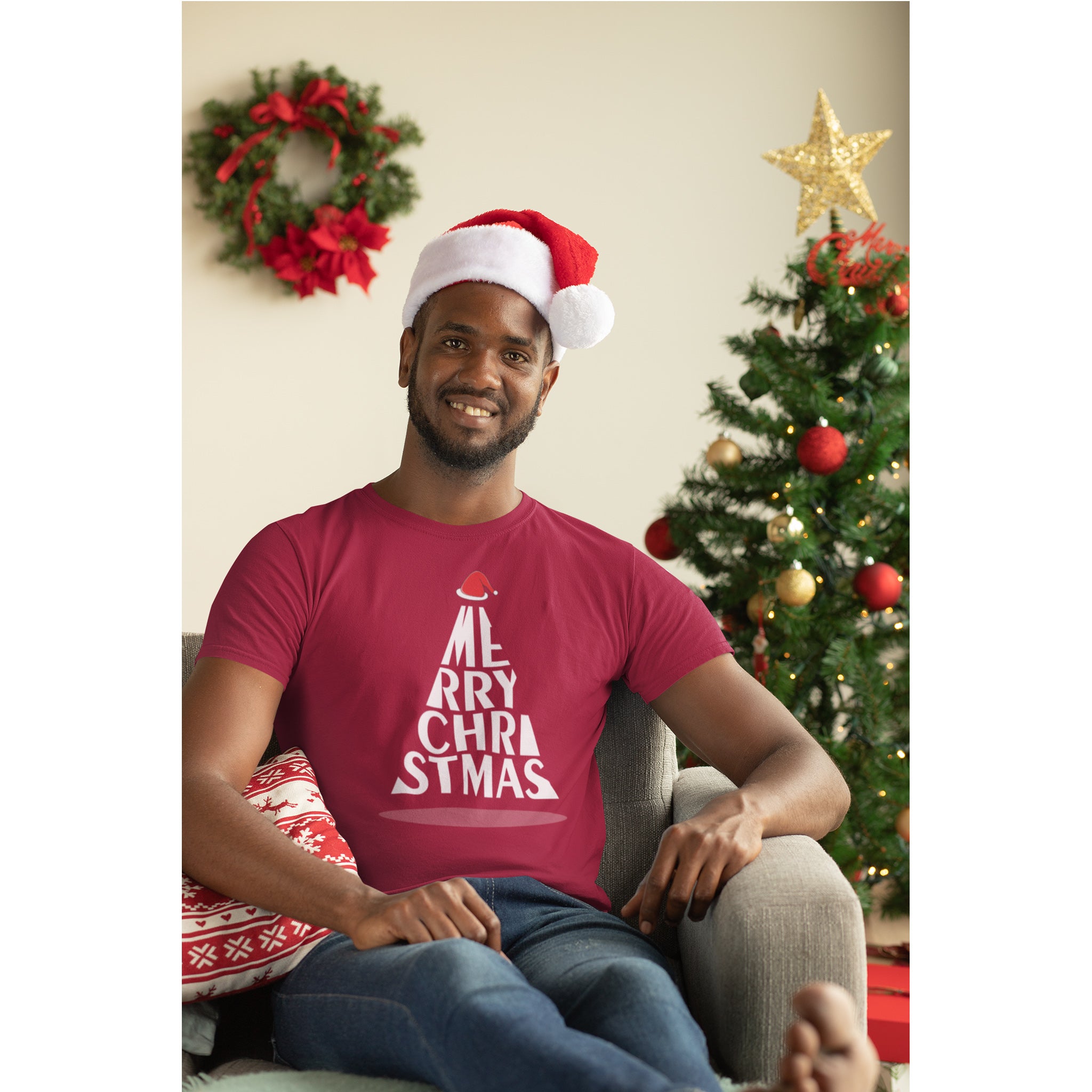Man wearing a red 'Merry Christmas' t-shirt and Santa hat in a festive living room.