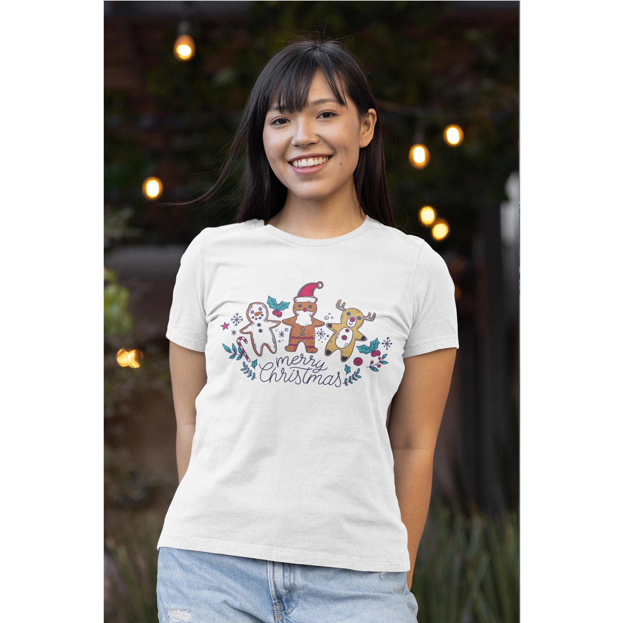 Woman wearing a white t-shirt with a Christmas design, standing outdoors with lights in the background.